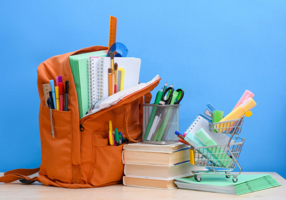 Orange school backpack full of school supplies and a supermarket basket with office supplies on a blue background.
